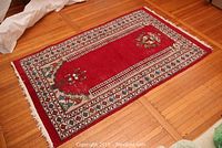 Full carpet view showing red background, white and multicolor borders, fringes on edges, placed on wooden floor.