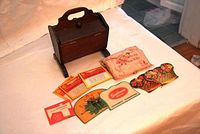 Wooden sewing basket with vintage embroidered and advertising needlebooks laid out in front on white cloth surface.