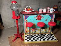 Full view of the Rock and Roll cafe counter with three red stools and the red candy dispenser beside it with basket of toy fruit and mugs on counter.