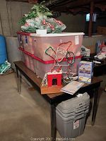 Full view of table with two red-lid plastic bins stacked, topped with Christmas greenery and floral arrangements. Light-up Santa decoration and box of mini lights next to bins. Plastic storage tote visible under table.