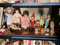 Wide view of shelf showing three 8-inch unmarked girl dolls, Coca Cola toy truck, several Wizard of Oz figurines including Pinocchio puppet, and Russian nesting dolls.