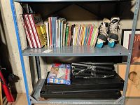 Metal shelf unit with Spanish course books, board games, ladies ice skates, disassembled plastic shelf unit parts on bottom shelf