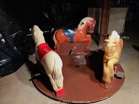 Photo of three plastic carousel horses mounted on a brown circular base, showing side view of cream, tan, and orange horses with pedals and handles