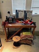 Wood tool bench with Craftsman bench vice, Craftsman bench grinder with lamp, Black and Decker hedge trimmer, two unopened Builders Edge gable vents, three unopened bags of cedar mulch, and wooden spool on floor.