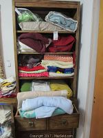 Wooden shelving unit with multiple folded blankets and throws assorted in colors including maroon, red, cream, yellow, green, blue; a few plastic bags at top shelf; blankets folded inside drawer at bottom.