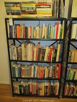 Photo showing several shelves filled with a large collection of books, both hard cover and paperback, arranged in rows on metal shelving units in a well-lit room.