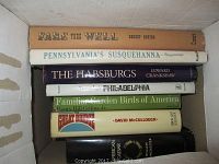 Books stacked horizontally inside a cardboard box showing titles on spine including 'Fare Thee Well', 'Pennsylvania's Susquehanna', 'The Habsburgs', 'The Great Bridge' and others.