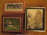Three framed artworks on carpeted floor: a framed print with a decorative border and stylized figures, an oil painting of a garden scene with flowers and rocks, and a framed landscape painting with trees and rocks.