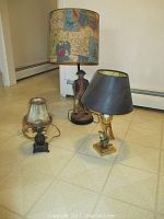 Three lamps set on a tiled floor: colonial figurine lamp with nautical map shade, horn-shaped lamp with black shade, pineapple-shaped lamp with fringed lampshade