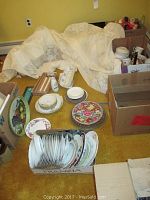 Overview image showing boxes and grouping of various plates, some stacked and some displayed on a table beside a lace fabric and other boxes.