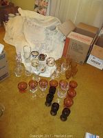 Collection of glassware, vases, small ceramic creamers, decanter, and various stemware grouped on a carpeted floor with boxes and fabric in the background.