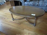 Wood oval coffee table showing top surface and legs, placed on a wood floor with couch and furniture in background.