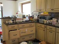 Overview of kitchen counter and cabinets with multiple kitchenware items piled on countertop including plastic containers, cups, a toaster oven and various other items