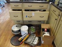 Wide view showing various pots, baking pans, measuring cup, rolling pin, and a metal colander along with a box of small kitchen items.