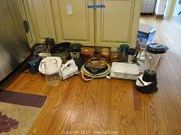 Several small kitchen appliances and bakeware arranged on wooden floor, including George Foreman grill, blender, coffee grinder, pans, and radio.