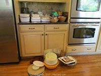 Wide view of kitchen showing Corningware collection stacked on countertop and floor in front of cabinets. Various baking dishes, bowls, and a basket visible.