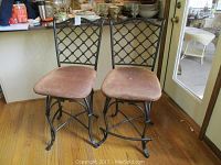 Front view of two swivel bar stools with lattice iron backs and fabric cushions, standing on wood floor near kitchen counter and door.