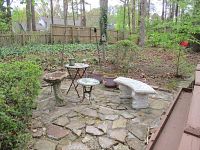 Wide view of outdoor stone patio area with cement bird bath, two mosaic iron tables, curved cement bench, and round planters.