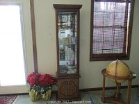 Full view of the curio cabinet standing next to a window and globe stand, showing wooden lower cabinet doors and glass display shelves with mirrored back.