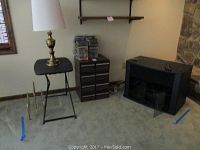 Room corner with brass lamp on small black folding table, four VCR storage cabinets stacked, black wood and smoked glass entertainment cabinet, and power cords on floor