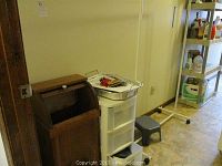 Photo showing a wood roll-top trash can, a white 3-drawer plastic storage cabinet, small step stool, and a metal rack with household bottles.