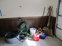 Photo of assorted garden tools including hoses, buckets, broom, and gas can clustered against wall