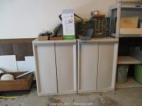 Two gray plastic cabinets side by side with double doors closed, located against wall in workshop.