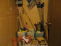 Photo showing corner storage filled with various yard tools including rakes, 4 garden torches, plant stands on small wheels, a basket, and an assortment of clay pots stacked on the floor.