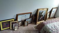 Photo showing a variety of framed pictures placed on the floor leaning against a wall next to a bed, shows different frame styles and art subjects including maritime ships, floral, portrait, and a monkey with palm trees.