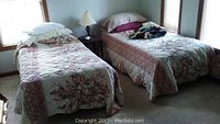 Two twin beds with floral bedspreads, pillows, quilts, a nightstand between them, a lamp, and clock radio on the nightstand
