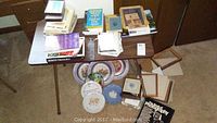 Overview of laminate table containing stacks of assorted books, papers, frames on top. Plates and additional plates placed underneath table.