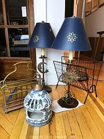 Photo of the pair of metal lamps, porcelain lantern, brass wired magazine racks all together on wooden floor.