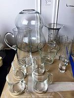 Clear glassware set including various glass items such as a large bulbous vase, pitcher, mugs, and drinking glasses arranged on table.