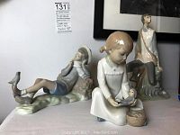 Wide shot showing all three porcelain figurines: a boy with bird by Lladro, NAO boy reclining with bird, and NAO girl standing.