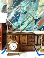 Wooden jewelry box with two doors and eight drawers, brass geometric sculpture on top, and brass arched clock in front, all displayed on a table with candlesticks.