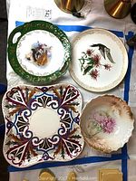 Four decorative plates and bowls displayed on table: Imperial Vienna Deco square platter, Boehm Studios hummingbird plate, Bavaria fruit bowl, Victoria courting scene platter.