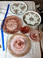 Top down view showing pink depression glass cake dish with central handle, footed candy dish, small opalescent vase, pink platter with gold floral border, and two porcelain platters with floral designs