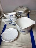 Full view of nine Corning Ware Cinderella bakeware pieces including rectangular dishes and large square dish with glass lid, plus three bone china saucers in front.