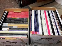 Two boxes filled with assorted art and design auction catalogs from various auction houses, showing spines with diverse logos and colors.