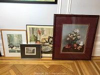 Four framed art prints leaning against wall, including street scenes, poster and still life