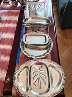 Photo of a collection of silver plate serving trays and chafing dish displayed on table, showing different shapes and sizes.