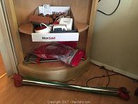 A corner shelf holding a green and red stepper on the floor, a pair of dumbbells, and some wooden hand grips.
