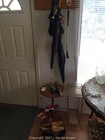 Wide view showing wood side table, hanging umbrellas, metal shoe horns, wooden plaques, wooden box, umbrella stand and glassware on table