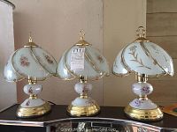 Three glass and brass table lamps with floral designs on white glass shades and bases, brass trim, set on a black surface against a neutral background.