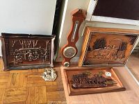 Photo of copper covered wagon scene carving, wooden barometer and two framed wooden train reliefs on floor against wall.