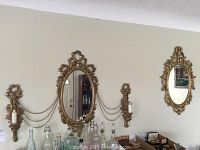 Two gold framed oval mirrors with matching gold wall candle sconces hung on wall. Chains connect sconces to mirrors. Frames appear to be plastic with metal sconces.