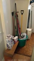 Photo showing multiple cleaning tools including brooms, mops, shovel, buckets, and a small table.
