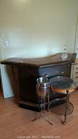 Photo showing side view of vintage rollable bar with dark brown vinyl upholstery and wood top, with wine chiller, decanter on top, and rolling glass-shelved serving tray in front.