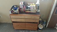 Wide view showing a wooden cabinet with drawers and various boxes on top including light bulbs and a white desk lamp, along with paint cans on the floor nearby.