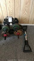 Dumbbells with multiple green and brown weight plates on a black stand, placed on carpeted floor against wood panel wall.